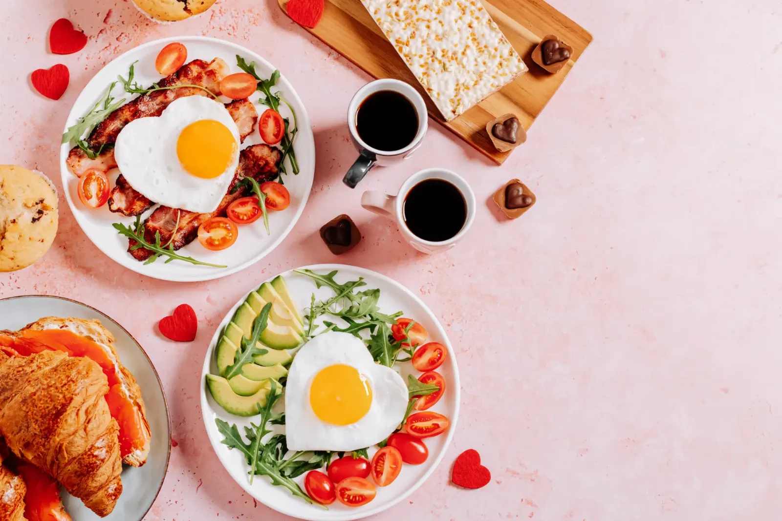 Brunch spread with heart-shaped fried eggs, bacon, avocado, croissants and coffee on a pink table