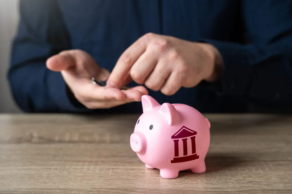 Hand dropping a coin into a pink piggy bank marked with a bank symbol