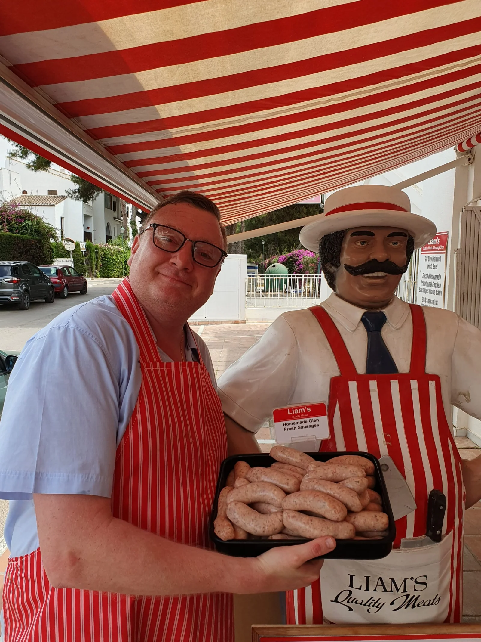 Liam at Liam's Quality Meats with a tray of their own sausages, beside the butcher statue