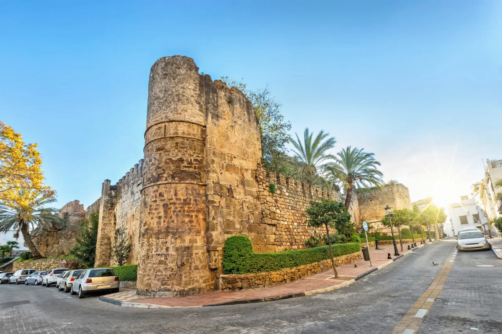 Old castle walls in Marbella's historic centre, lined with palm trees and parked cars