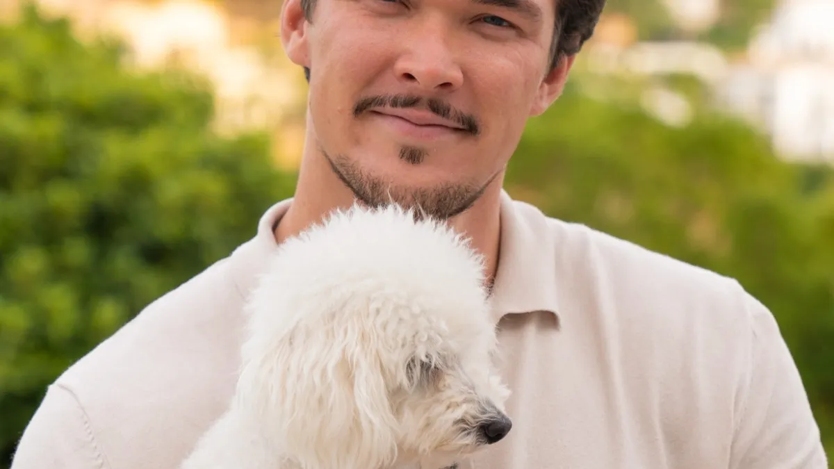 Smiling man holding a small white fluffy dog outdoors with greenery behind