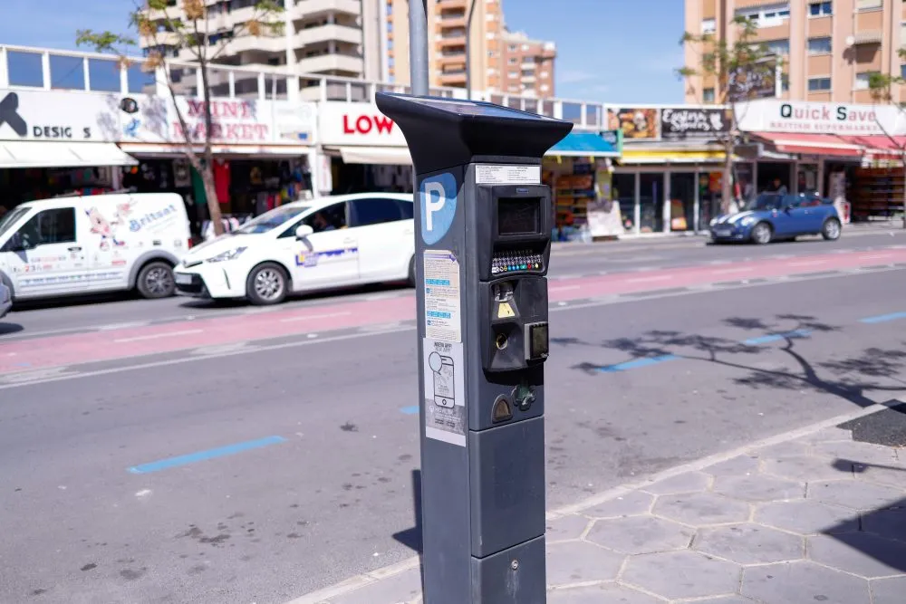 Solar-powered parking meter on a street with blue-line parking bays and shops behind