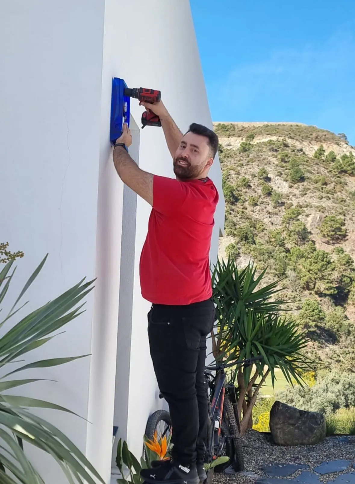 Un t&eacute;cnico con camiseta roja taladrando para instalar una alarma en la pared exterior blanca de una villa